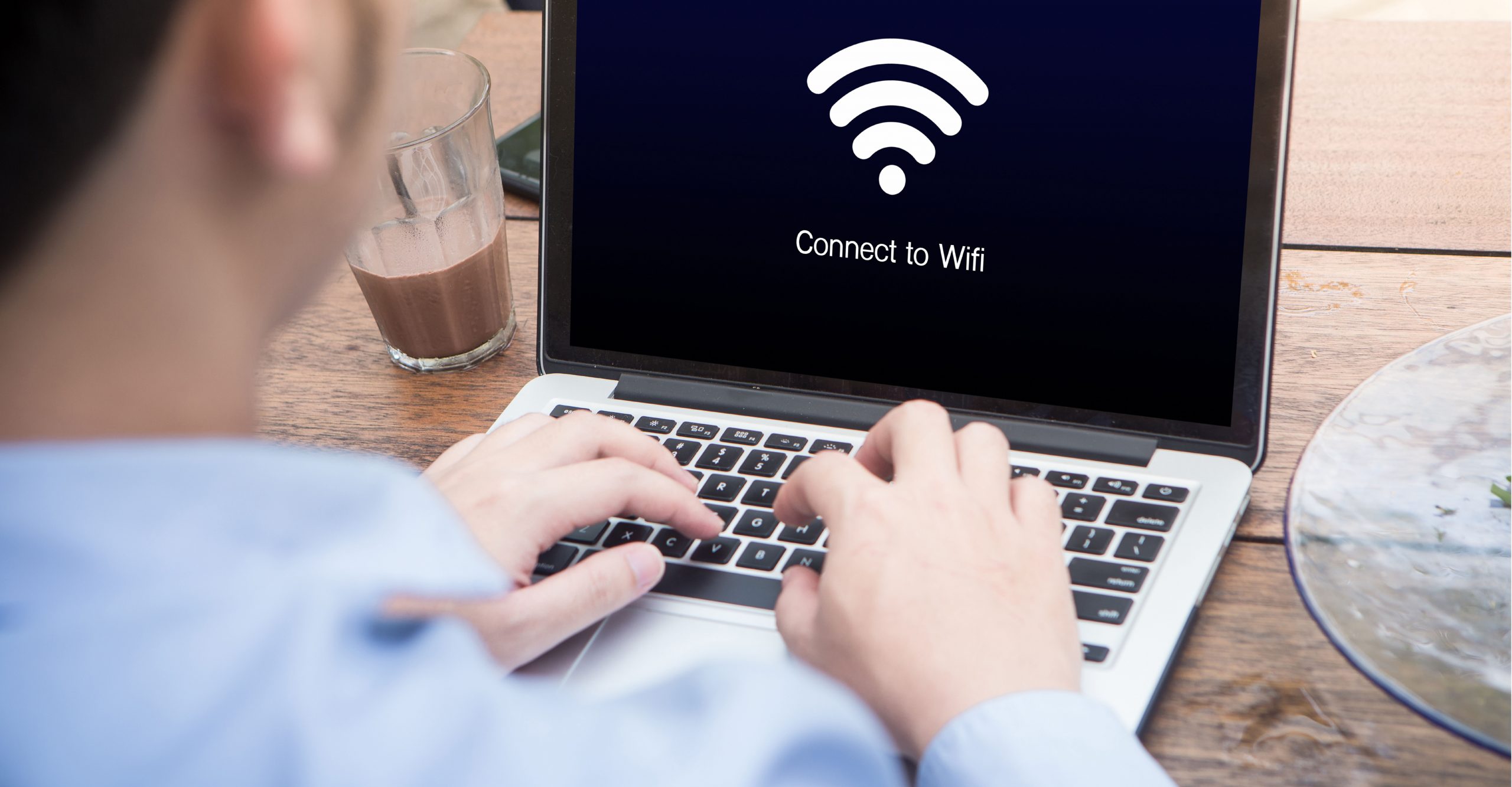 Person typing on a laptop displaying a "Connect to Wifi" message while sipping a glass of chocolate drink on the wooden table beside it, ensuring network uptime.