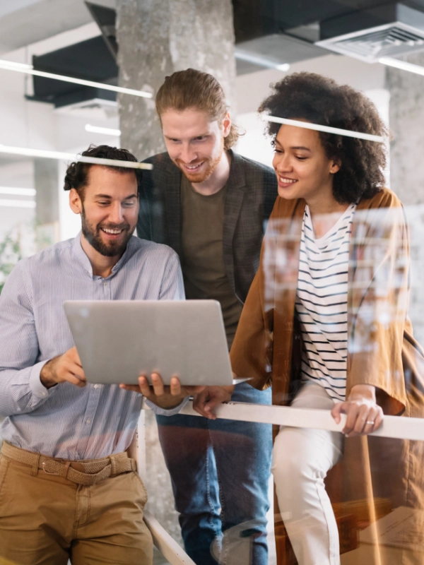 Three people standing together, looking at a laptop screen and smiling in a modern office setting of an IT support company.