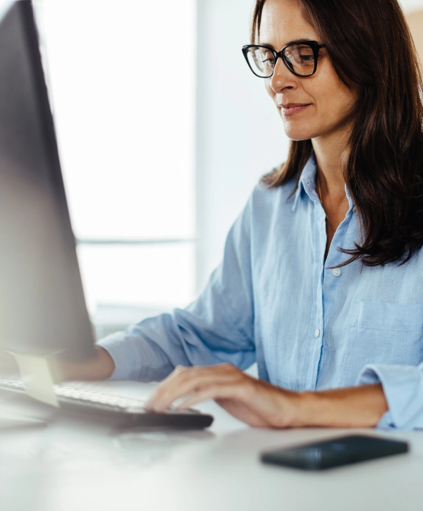 A woman wearing glasses and a blue shirt is seated at a desk, typing on a keyboard with a computer monitor in front of her, working for an IT support company.