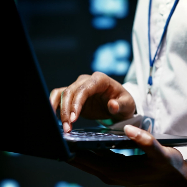 Close-up of a person typing on a laptop keyboard, with a faintly visible screen in the background. The individual, wearing a white shirt and a lanyard, appears intent on their task, likely working for an IT support company.