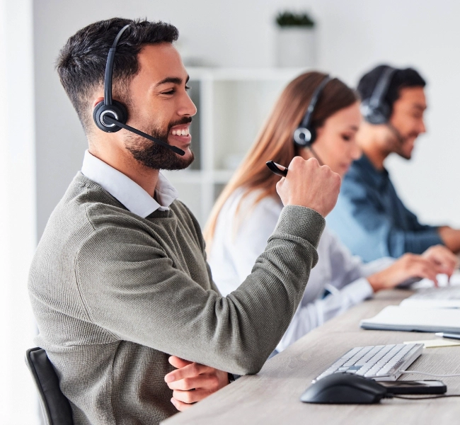 Three call center employees wearing headsets work at their desks. The man in the foreground is smiling and holding a pen, showcasing their dedication to providing IT services. A woman and another man are working on computers in the background, ensuring smooth network support for clients.