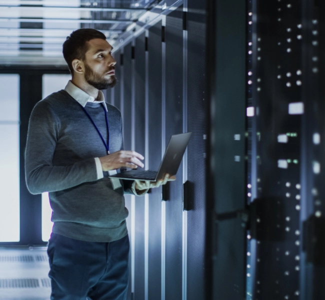 A man holding a laptop inspects server racks in a data center, ensuring the highest quality of managed IT services.