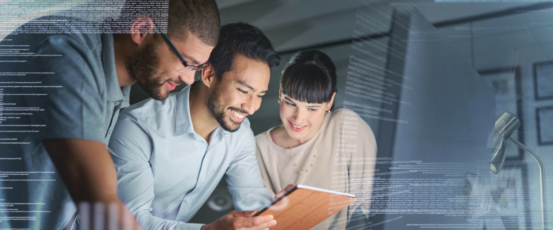 Three people are smiling and looking at a tablet together in a dimly lit room, with lines of code superimposed in the foreground, epitomizing the essence of computer IT support.