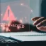 An image of a person's hand hovering over their keyboard while an abstract image saying "System Hacked" emulates from the computer screen.