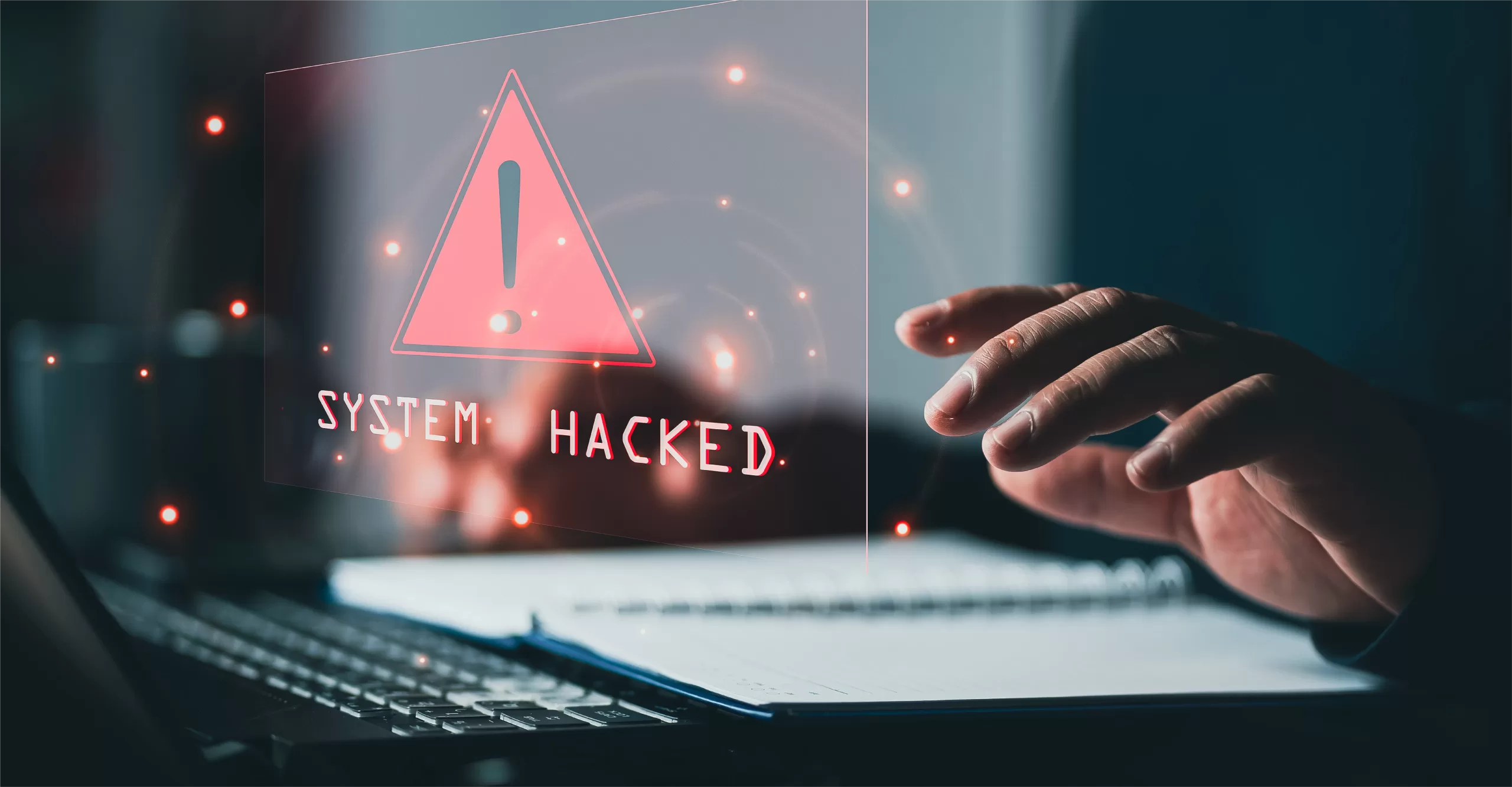 An image of a person's hand hovering over their keyboard while an abstract image saying "System Hacked" emulates from the computer screen.